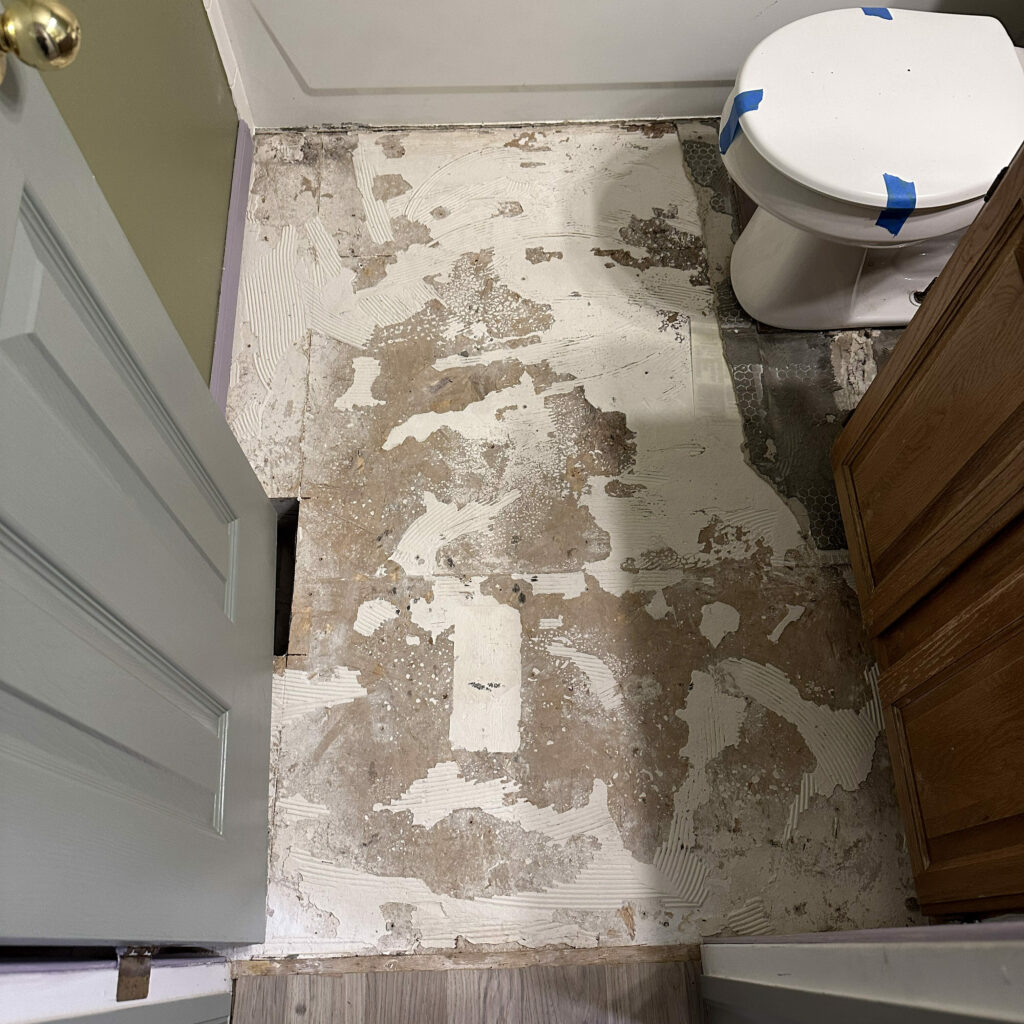 Pictured: an exposed subfloor in a small bathroom with bits of white thinset covering parts of it after removing old floor tile.