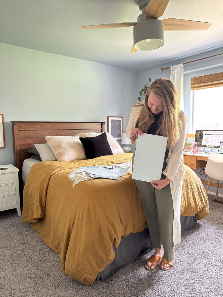 woman holding a light blue peel and stick paint sample in a blue bedroom with a yellow bedspread.