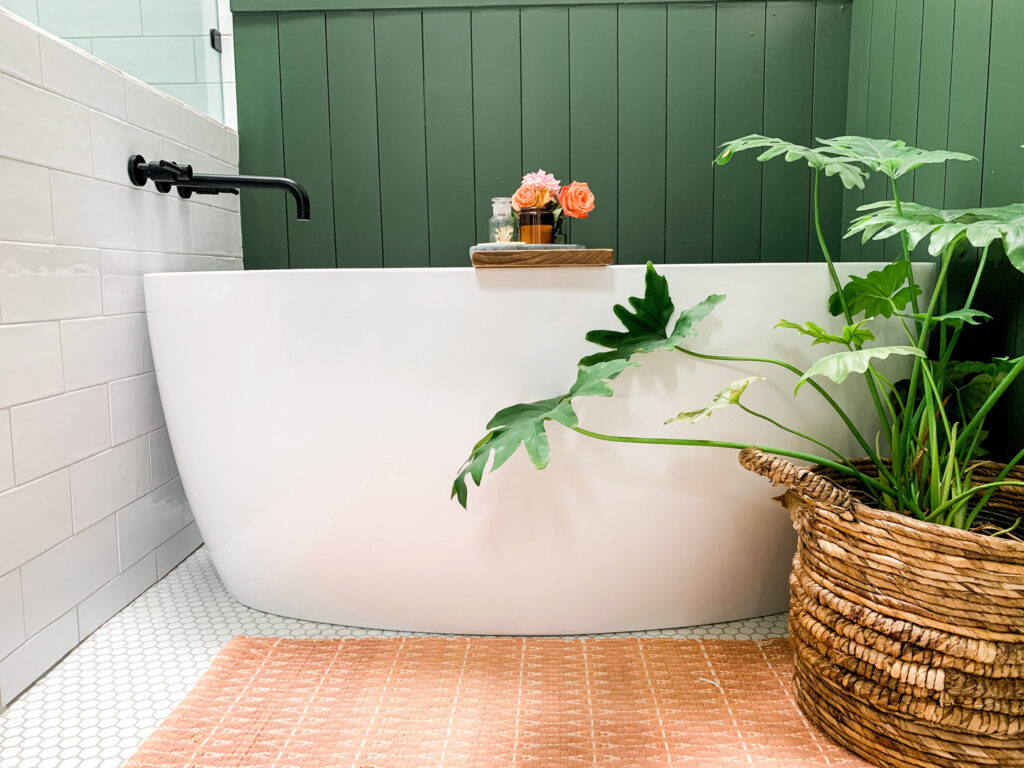 Freestanding tub on a white hex tile floor with a green shiplap background, rust orange bath mat and large plant in a basket